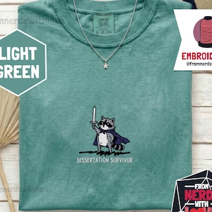 May include: A light green t-shirt featuring an embroidered graphic of a raccoon wearing a purple cape and holding a sword. Below the raccoon, the text reads DISSERTATION SURVIVOR. The shirt has a crew neckline and a silver star pendant necklace.