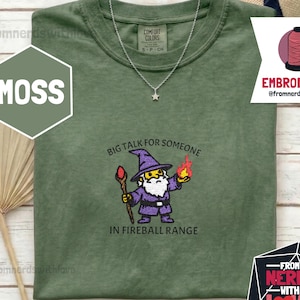 May include: Moss green t-shirt with a wizard graphic and the text "BIG TALK FOR SOMEONE IN FIREBALL RANGE." The shirt has a crew neck and short sleeves. A silver necklace with a star pendant is visible. The shirt is from the brand Comfort Colors.