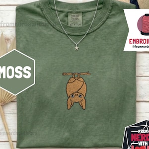 May include: A moss green t-shirt featuring an embroidered bat hanging upside down from a branch. The shirt has a crew neck and short sleeves. A silver necklace with a star pendant is visible. The text "MOSS" is in a hexagon shape.