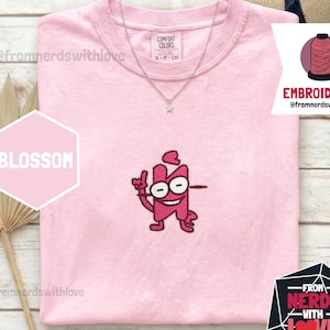 May include: A light pink Comfort Colors t-shirt with an embroidered cartoon character design. The design is a pink figure with large eyes and a smiling face. The shirt also features the text "BLOSSOM" and "EMBROIDERED". A silver necklace with a star pendant is visible.