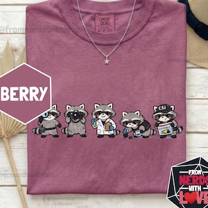 May include: A berry-coloured Comfort Colors t-shirt with a row of cartoon raccoons in different roles, such as a photographer and a CSI agent. The shirt features the text "BERRY" and "FROM NERDS WITH LOVE". A silver necklace with a star pendant is also visible.