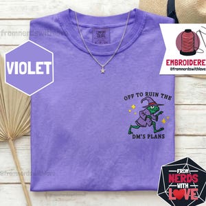 Może przedstawiać: Fioletowa koszulka Comfort Colors ze srebrnym naszyjnikiem w kształcie gwiazdy. Koszulka posiada haftowany wzór zielonej żaby czarodzieja z napisem "OFF TO RUIN THE DM'S PLANS". Logo z napisem "EMBROIDERED" znajduje się w prawym górnym rogu.