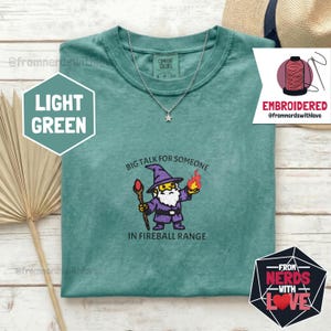 May include: Light green t-shirt with a wizard graphic and the text "BIG TALK FOR SOMEONE IN FIREBALL RANGE." The shirt also features a small star pendant necklace. The shirt is part of the "Comfort Colors" collection.