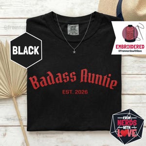 May include: Black t-shirt with the text "Badass Auntie EST. 2026" in red gothic font. A silver necklace with a star pendant is visible. The shirt has a crew neck and short sleeves. The word "BLACK" is in a black hexagon.