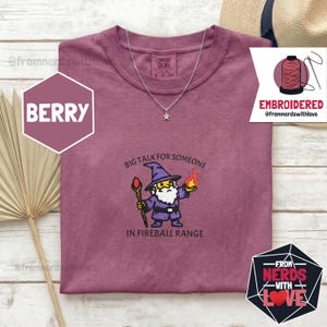 May include: Berry-colored t-shirt with an embroidered wizard design. The wizard holds a staff and a fireball, with the text "BIG TALK FOR SOMEONE IN FIREBALL RANGE." A silver necklace with a star pendant is also visible.
