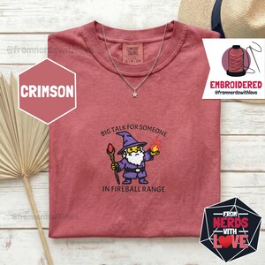May include: Crimson-colored t-shirt with a wizard graphic and the text "BIG TALK FOR SOMEONE IN FIREBALL RANGE." The shirt also features the text "EMBROIDERED @fromnerdswithlove" and "-FROM NERDS WITH LOVE-". A silver necklace with a star pendant is on the shirt.