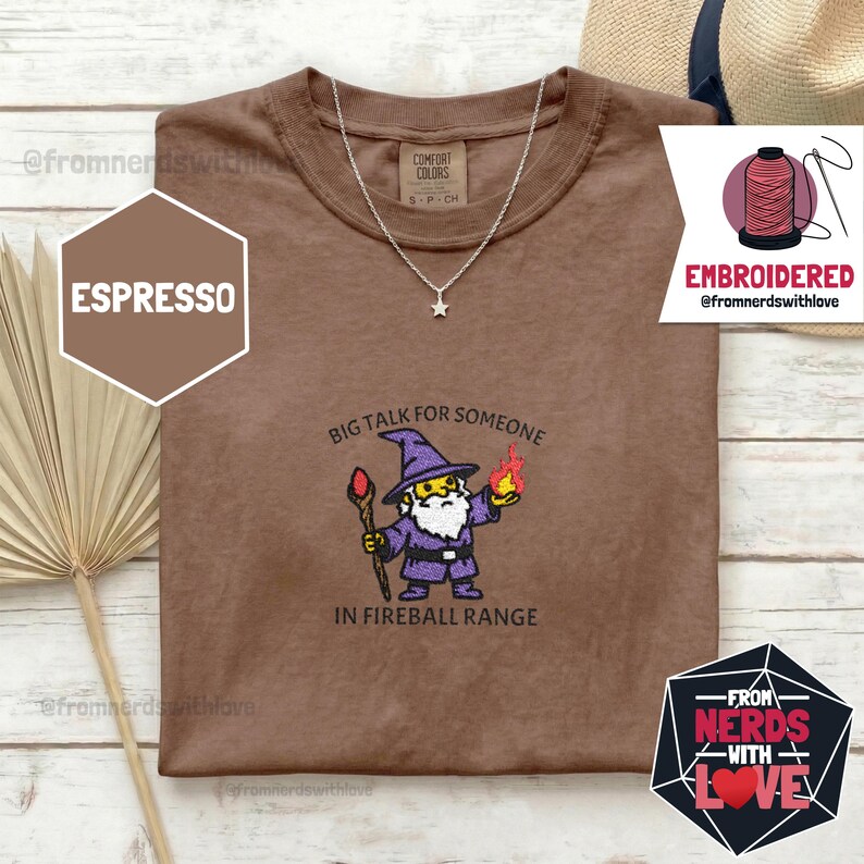 May include: Espresso-colored t-shirt with an embroidered wizard graphic. The wizard is depicted in purple robes, holding a staff and a fireball, with the text "BIG TALK FOR SOMEONE IN FIREBALL RANGE". A silver star pendant necklace is also visible.