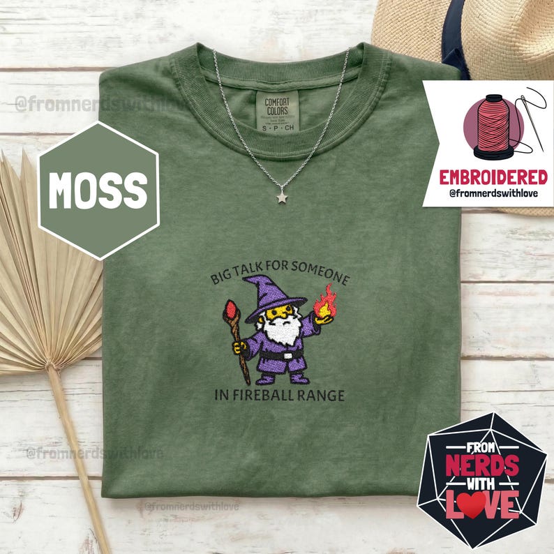 May include: Moss green t-shirt with a wizard graphic and the text "BIG TALK FOR SOMEONE IN FIREBALL RANGE." The shirt has a crew neck and short sleeves. A silver necklace with a star pendant is visible. The shirt is from the brand Comfort Colors.