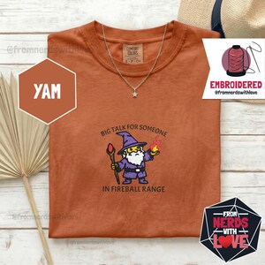 May include: Rust-colored t-shirt with a wizard graphic and the text "Big Talk for Someone in Fireball Range." The shirt also features a silver necklace with a star pendant. The shirt is a Comfort Colors brand.
