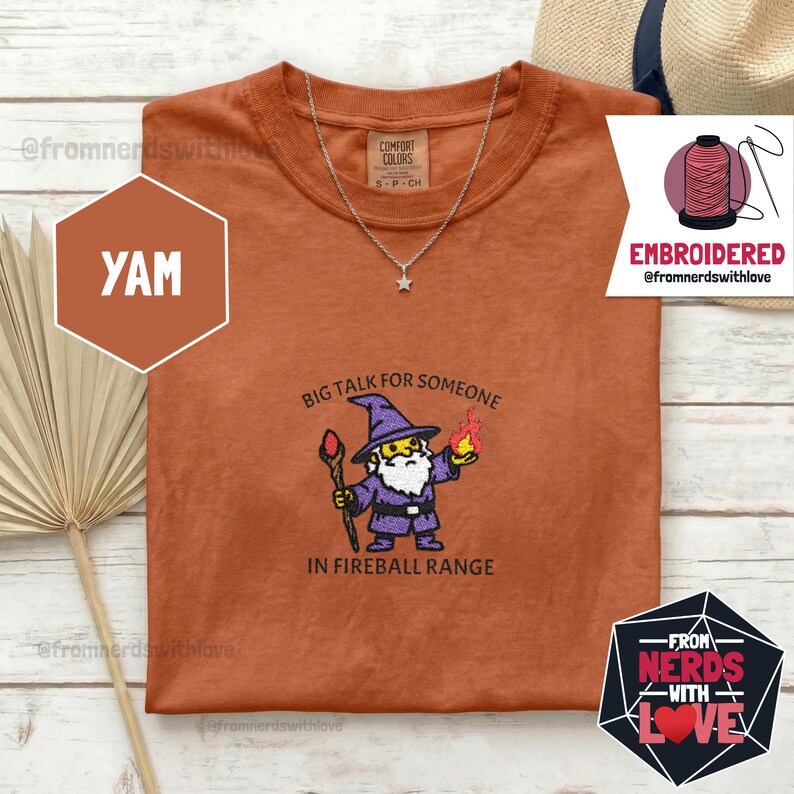 May include: Rust-colored t-shirt with a wizard graphic and the text "Big Talk for Someone in Fireball Range." The shirt also features a silver necklace with a star pendant. The shirt is a Comfort Colors brand.