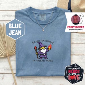 May include: Blue jean Comfort Colors t-shirt with an embroidered wizard graphic. The wizard holds a staff and a fireball, with the text "BIG TALK FOR SOMEONE IN FIREBALL RANGE". A silver necklace with a star pendant is also visible.