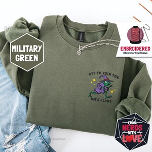 Może przedstawiać: Zielona bluza w kolorze wojskowym z haftowanym napisem "OFF TO RUIN THE DM'S PLANS" i żabą w stroju czarodzieja. Widoczny jest srebrny naszyjnik. Bluza jest złożona na niebieskich dżinsach.