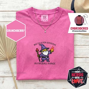 May include: A pink t-shirt with an embroidered wizard graphic holding a staff and fire, with the text "BIG TALK FOR SOMEONE IN FIREBALL RANGE." A silver necklace and a hat are visible. The shirt has the text "CRUNCHBERRY" and "EMBROIDERED".