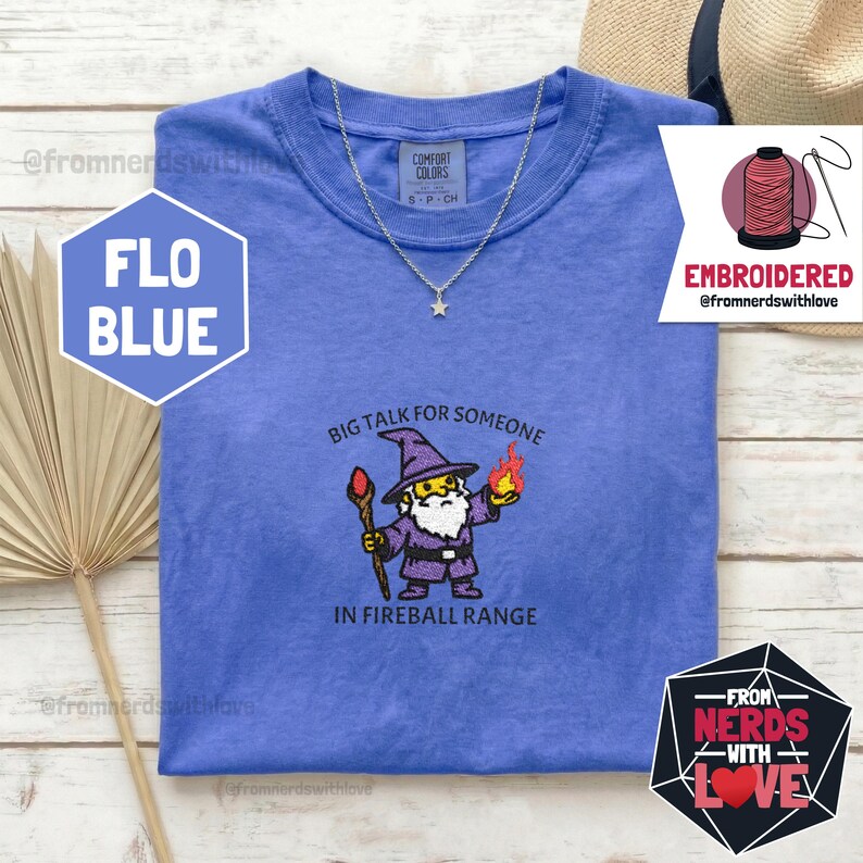 May include: A blue t-shirt with a wizard graphic holding a staff and a fireball, with the text "BIG TALK FOR SOMEONE IN FIREBALL RANGE." A silver necklace with a star pendant is on the shirt. The shirt is labeled "FLO BLUE."