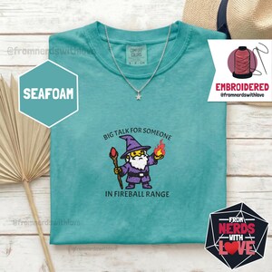 May include: Seafoam green t-shirt with a wizard graphic holding a staff and a fireball, with the text "BIG TALK FOR SOMEONE IN FIREBALL RANGE." A silver necklace with a star pendant is also visible.