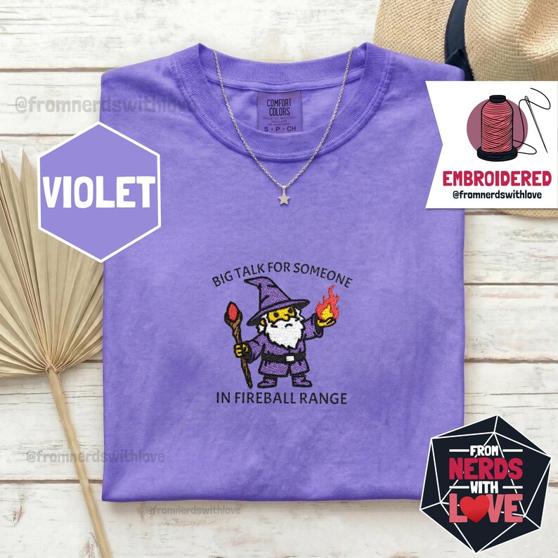 May include: Violet Comfort Colors t-shirt with an embroidered wizard graphic. The wizard holds a staff and a fireball, with the text "BIG TALK FOR SOMEONE IN FIREBALL RANGE." A silver star necklace is also visible.