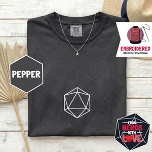 May include: A dark gray t-shirt with a white geometric design and a star pendant necklace. The shirt has the word "PEPPER" in a white font on a dark gray hexagon. The shirt also has the text "EMBROIDERED" and "FROM NERDS WITH LOVE".