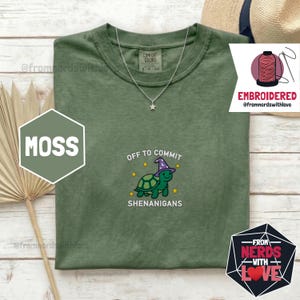 Könnte beinhalten: Ein moosgrünes T-Shirt mit einem gestickten Design mit einer Schildkröte mit Hexenhut, dem Text "OFF TO COMMIT SHENANIGANS" und einer Sternkette. Das Shirt hat auch den Text "COMFORT COLORS" auf dem Etikett.