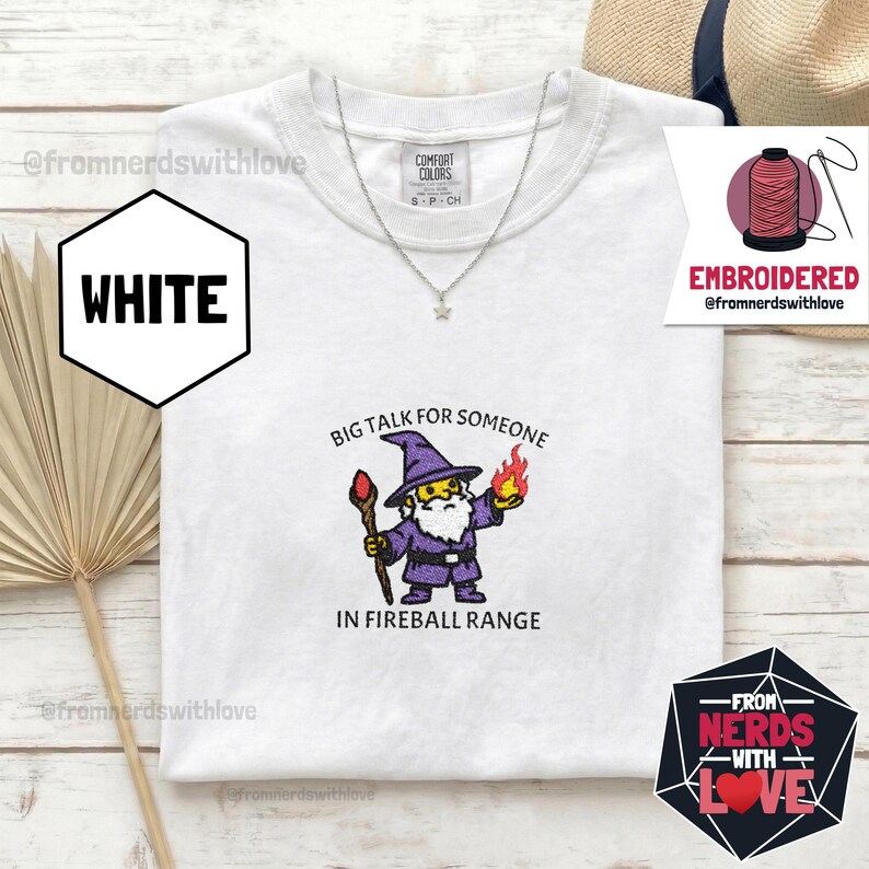 May include: White t-shirt with an embroidered wizard graphic. The wizard is depicted in purple robes, holding a staff and a fireball, with the text "BIG TALK FOR SOMEONE IN FIREBALL RANGE". A silver necklace is visible.