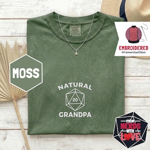 May include: A moss green t-shirt with the words "NATURAL GRANDPA" and a geometric design. A silver necklace with a star pendant is visible. The shirt also has the text "COMFORT COLORS" and "EMBROIDERED @fromnerdswithlove".