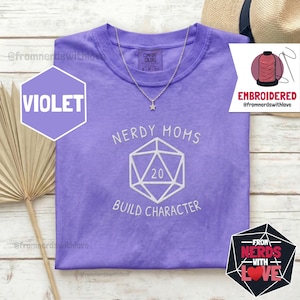 May include: A violet short-sleeved t-shirt featuring white embroidery of a twenty-sided die with the number 20 in the centre. The text NERDY MOMS is arched above the die, and the text BUILD CHARACTER is arched below the die on the chest.