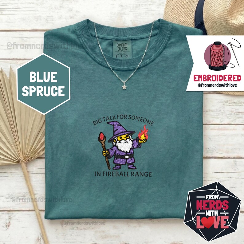 May include: Blue spruce t-shirt with a wizard graphic holding a staff and a fireball, with the text "BIG TALK FOR SOMEONE IN FIREBALL RANGE." The shirt also has a silver necklace with a star pendant.