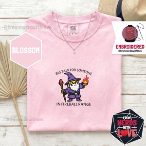 May include: Pink t-shirt with the embroidered phrase "BIG TALK FOR SOMEONE IN FIREBALL RANGE" and a cartoon wizard design. The shirt has a crew neck and short sleeves. A silver necklace with a star pendant is visible.