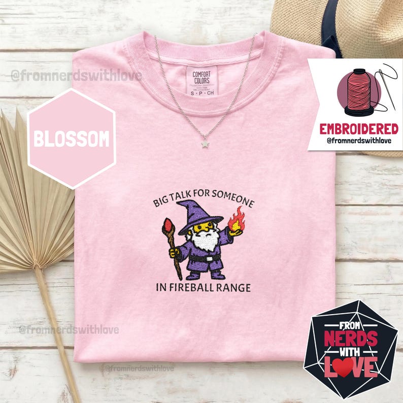 May include: Pink t-shirt with the embroidered phrase "BIG TALK FOR SOMEONE IN FIREBALL RANGE" and a cartoon wizard design. The shirt has a crew neck and short sleeves. A silver necklace with a star pendant is visible.