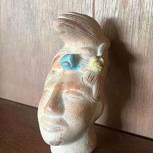 May include: A terracotta-colored clay sculpture of a human head featuring an elaborate headdress. The figure has a smooth, matte finish with small, distinct blue and yellow painted accents on the forehead area and carved linear details.