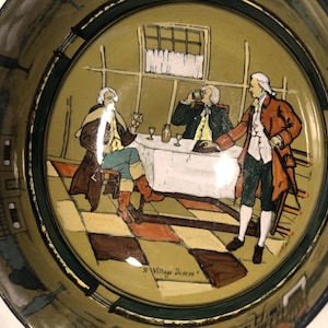 May include: A ceramic bowl with a green and brown rim and a painted scene of three men in a room with a checkered floor. The scene includes a table with a white tablecloth and a bottle of wine. The bowl is inscribed with the text "Village Scene".