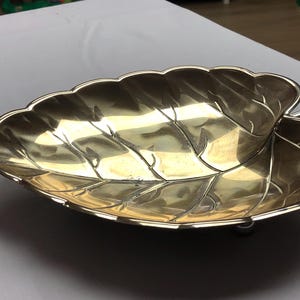 Può includere: Un piatto da portata in argento a forma di foglia con un design in rilievo. Il piatto ha un manico su un lato e tre piedini sul fondo.