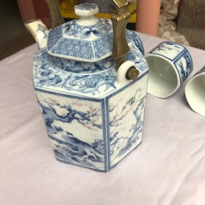 Peut inclure: Une théière en porcelaine bleue et blanche avec une poignée et un couvercle en laiton. La théière est décorée d'un motif floral et repose sur une surface blanche.