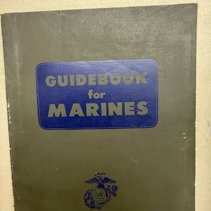 May include: A vintage guidebook for Marines. The cover is a muted olive green with a blue rectangle containing the words "GUIDEBOOK for MARINES" in bold, sans-serif font. A blue emblem is at the bottom.