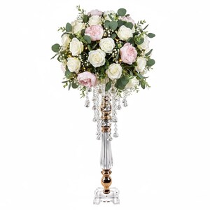 Puede incluir: Un centro de mesa alto con un arreglo esf&eacute;rico de rosas blancas y rosa p&aacute;lido, hojas de eucalipto verdes y paniculata. La bola floral se asienta sobre un soporte de vidrio estriado transparente con detalles dorados y cuentas de cristal.