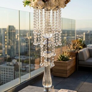 Puede incluir: Un centro de mesa alto de acr&iacute;lico transparente y metal plateado con un dise&ntilde;o escalonado y cuentas colgantes facetadas tipo cristal. La parte superior sostiene un gran arreglo floral redondeado de flores artificiales color crema y rosa viejo.