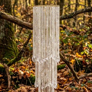 May include: A three-tiered cylindrical chandelier featuring numerous strands of clear, faceted crystal beads suspended from a circular gold-toned frame. The structure hangs from a gold-toned hook and includes pointed crystal drops at the base of each tier.