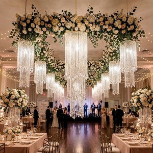 May include: A large indoor event space featuring multiple tiered crystal chandeliers hanging from the ceiling, surrounded by extensive floral arrangements of white roses and greenery, with string lights draped across the room.