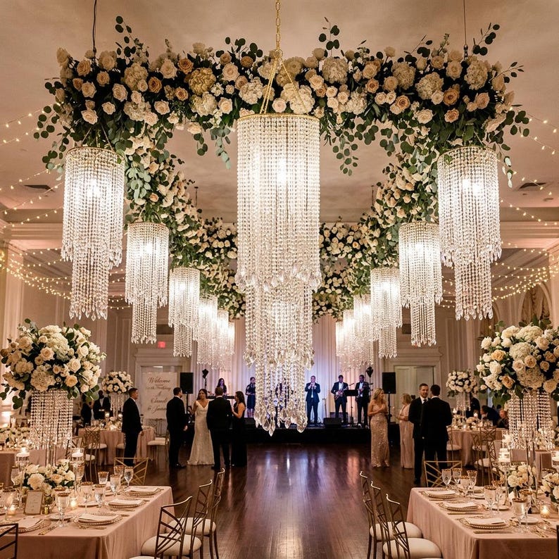 May include: A large indoor event space featuring multiple tiered crystal chandeliers hanging from the ceiling, surrounded by extensive floral arrangements of white roses and greenery, with string lights draped across the room.
