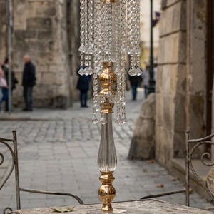 Puede incluir: Un soporte decorativo alto para centro de mesa con base y tallo de metal dorado y detalles en acr&iacute;lico transparente. El marco circular superior est&aacute; adornado con m&uacute;ltiples hileras de cuentas transparentes colgantes tipo cristal.