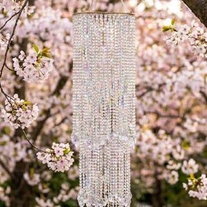 May include: A tiered crystal chandelier wind chime featuring multiple strands of clear, faceted glass beads hanging from a circular frame. The structure is suspended by a clear plastic loop and features varying lengths of beads ending in pointed crystals.