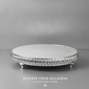 May include: A round silver cake stand with a mirrored top surface and a decorative embossed border featuring a repeating pattern. The stand is supported by three small, curved feet. The text "ELEVATE YOUR OCCASION LUXURY CAKE STAND" is visible.