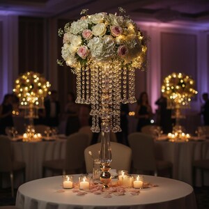 Puede incluir: Un alto soporte de centro de mesa de cristal transparente con base y tallo dorados, coronado por un gran arreglo floral de rosas y hortensias blancas y rosas, acentuado por hileras de cuentas de cristal colgantes y luces c&aacute;lidas integradas.