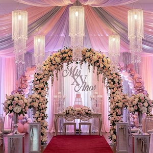 May include: A decorative event archway adorned with cream and pink roses, featuring a central sign reading Mis XV A&ntilde;os. The display includes crystal chandeliers, draped pastel fabric, balloon clusters, and a tiered cake on a table with a red carpet.