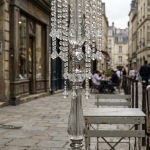 Puede incluir: Un centro de mesa alto de metal plateado y acr&iacute;lico transparente con un borde superior circular adornado con hileras colgantes de cuentas facetadas similares al cristal. La columna central tiene un dise&ntilde;o estriado sobre una base cuadrada.