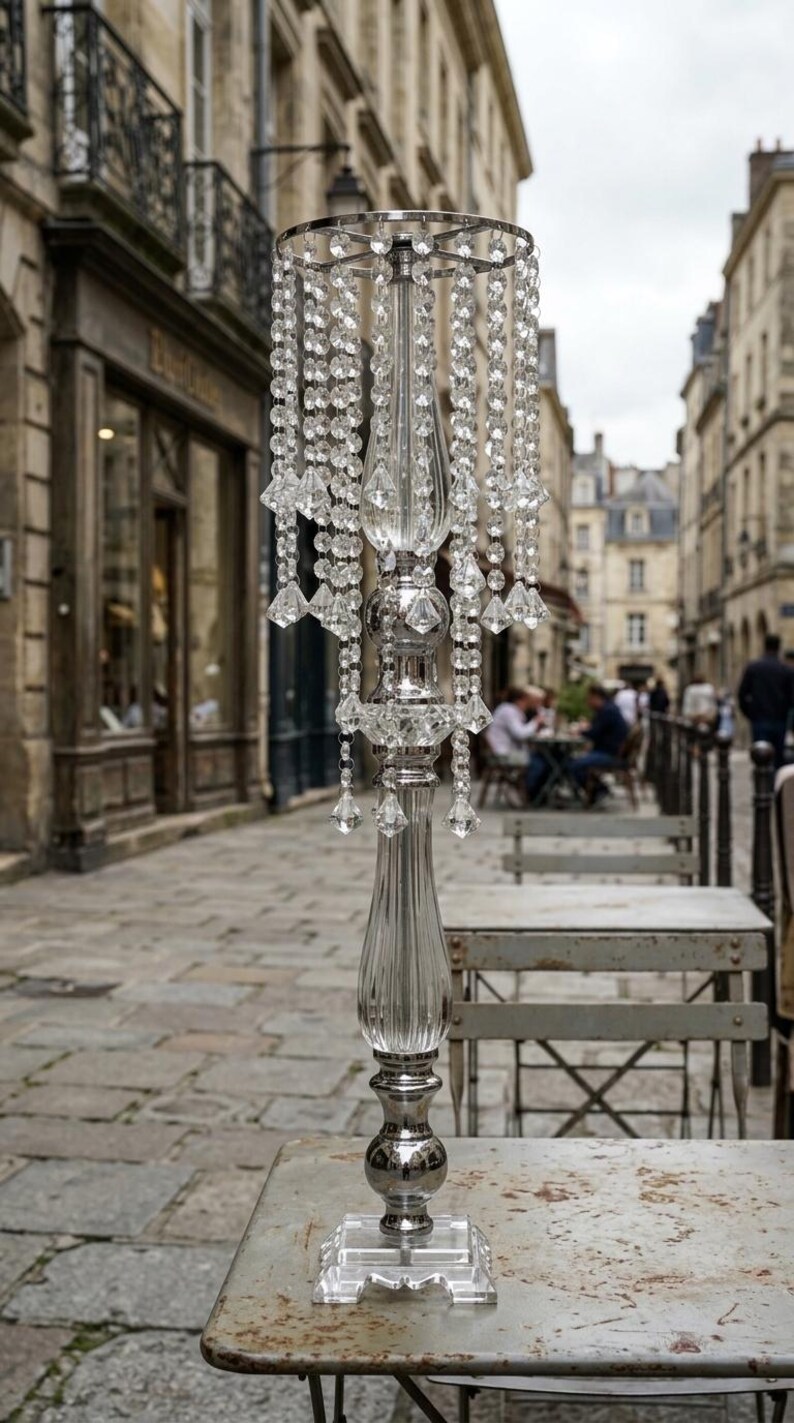 Puede incluir: Un centro de mesa alto de metal plateado y acr&iacute;lico transparente con un borde superior circular adornado con hileras colgantes de cuentas facetadas similares al cristal. La columna central tiene un dise&ntilde;o estriado sobre una base cuadrada.