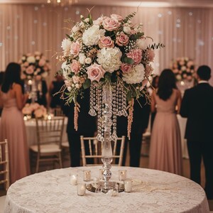 Puede incluir: Un alto soporte de centro de mesa de cristal transparente con cuentas colgantes tipo cristal y un gran arreglo floral de rosas rosas y blancas, hortensias blancas y follaje verde, sobre una mesa redonda con un mantel de encaje blanco.