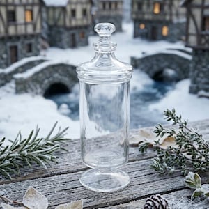 May include: A clear glass apothecary jar with a matching lid, featuring a cylindrical body, a pedestal base, and a small knob handle on top. The jar is transparent, smooth, and reflective, showcasing a simple design with clean lines and a polished finish.
