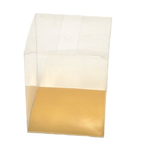 May include: A clear plastic box with a gold base. The box is empty and has a square shape.
