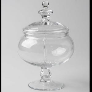 May include: A clear glass apothecary jar with a wide, rounded bowl, a pedestal base, and a matching removable lid featuring a spherical finial handle. The container has a smooth, transparent surface and a vertical seam along the center.