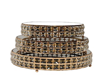 May include: A tiered cake stand with three circular levels. Each tier has a mirrored surface and is framed with gold-colored metal and square-cut crystal accents. The stand is designed for displaying desserts or decorative items.